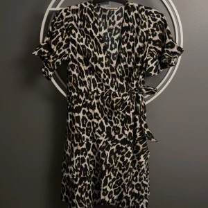 Leopardklänning Pernilla Wahlgren S - Snygg kort omlottklänning från Pernilla Wahlgren i leopardmönster. Klänningen har volangdetaljer, knyt i midjan och korta ärmar. Färgerna går i svart, beige och brunt. Perfekt för dig som vill sticka ut med en trendig look. Säljer då jag växt ur den, inte använd jättemånga gånger men fin till lite speciellare tillfällen eller bara vardagligt. 