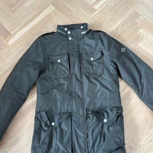 Svart parkajacka från J.Lindeberg XS - Snygg svart parkajacka från J.Lindeberg med fyra stora fickor framtill, hög krage och axelklaffar. Jackan har dragkedja och tryckknappar, samt en patch med logga på ärmen. Perfekt för dig som gillar stilren och funktionell design.