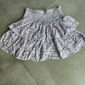 Blommig volangkjol med smock, stl 146-152 - Supersöt kort volangkjol med smock i midjan och blommigt mönster i lila, rosa, grön och vit. Kjolen har flera lager volanger som ger en fluffig och lekfull look. Perfekt för vår och sommar när du vill ha något lätt och färgglatt.