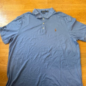 Blå pikétröja från Polo Ralph Lauren - Klassisk ljusblå pikétröja från Polo Ralph Lauren i bomull. Tröjan har krage, tre knappar och den ikoniska broderade polospelaren på bröstet. Classic fit-modell som är perfekt för en avslappnad men ändå stilren look.