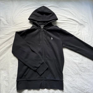 Svart hoodie från Polo Ralph Lauren XS - Svart hoodie från Polo Ralph Lauren i storlek XS. Klassisk design med huva, dragkedja och känguruficka framtill. Broderad vit logga på bröstet. Tillverkad i mjuk bomull, perfekt för chill dagar eller när du vill ha en clean streetstyle. Nypris på varan ligger på 2000kr i genomsnitt.