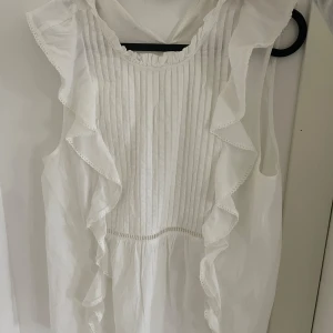 Vit volangblus med ärmlös design - Supersöt vit blus med volanger längs axlarna och ärmlös passform. Framsidan har smala veck och volangdetaljer som ger en romantisk vibe. Perfekt för dig som gillar en luftig och feminin stil. Blusen är gjord i ett lätt tyg och har rund halsringning.