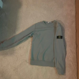  Stone Island sweatshirt - Säljer en grön-grå stilren sweatshirt från Stone Island Junior med klassisk patch på vänster ärm. Tröjan har rund halsringning och långa ärmar, perfekt för dig som gillar streetwear och enkel design. Materialet är mjukt och skönt, troligtvis bomull.