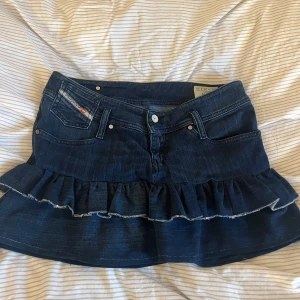 Mörkblå jeanskjol med volang - Säljer en mörkblå jeanskjol från Diesel med volangkant nedtill. Kjolen har klassiska jeansfickor fram och bak, bälteshällor och en snygg, lite ruffad volang som ger extra edge. Perfekt för dig som gillar denim och vill sticka ut lite extra.