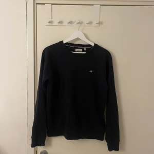 Mörkblå sweatshirt från Gant - Klassisk mörkblå sweatshirt från Gant med rund halsringning och broderad logga på bröstet. Tröjan har långa ärmar och ribbade muddar vid hals, ärmslut och nederkant. Perfekt för en stilren och avslappnad look. Pris kan diskuteras 