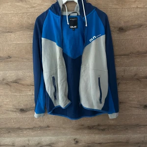Blå och grå vindjacka från CLN - Snygg vindjacka från CLN Athletics i blått och grått med huva och dragsko. Jackan har hel dragkedja framtill, två fickor med dragkedja och sportig design. Perfekt för dig som gillar en aktiv stil och vill ha något bekvämt och funktionellt.