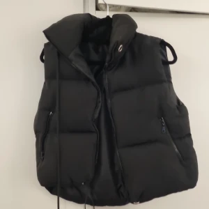 Svart dunväst med dragkedja, stl M - Säljer en svart, quiltad dunväst i storlek M. Västen har hög krage, två fickor med dragkedja och justerbar dragsko nertill. Tillverkad i 100% polyester och perfekt för lager-på-lager-stil under kyliga dagar. Märke okänt.