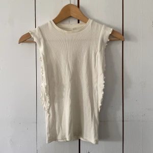 Vit ribbad ärmlös topp från Name It - Supersöt vit ribbad topp från Name It i ärmlös modell med volangkant längs ärmhålen. Toppens material är mjuk bomull och den har en tight passform som sitter snyggt. Perfekt för en fräsch och enkel look.