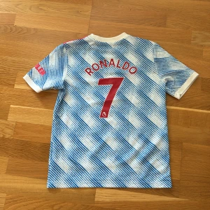 Manchester United Ronaldo bortatröja - Säljer en Manchester United bortatröja med Ronaldo 7 på ryggen. Tröjan är kortärmad och har ett blått och vitt randigt mönster, röda detaljer och klubbmärke framtill. Adidas-logga och sponsortryck. Perfekt för fotbollsfans!