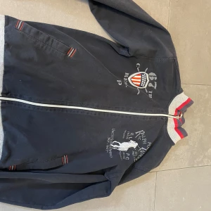 Tröja Ralph Lauren - Snygg mörkblå bomberjacka från Ralph Lauren med broderade detaljer och logga på bröstet. Jackan har grå ribbade muddar med röda och blå ränder, dragkedja framtill och två fickor. Perfekt för dig som gillar sportig och klassisk stil. Storlek 164-170 cm