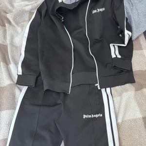 Palm Angels svarta mjukisbyxor - Svarta mjukisbyxor från Palm Angels med vita sidoränder och logga tryckt på ena benet. Byxorna har en sportig vibe och är perfekta för chill eller streetwear. Matchar med tillhörande zip-jacka för en komplett look.