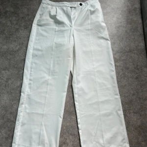 Vita vida kostymbyxor i satinlook - Snygga vita kostymbyxor med vida ben och hög midja. Byxorna har pressveck, sidfickor och stängs med både vit och svart knapp. Materialet har en lätt glansig satinlook som ger en clean och fräsch vibe.