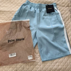 Palm Angels ljusblå shorts med vit stripe - Palm Angels shorts i ljusblått med vit sidoremsa och broderad logga på benet. Elastisk midja och sportig look, perfekta för sommaren. Snygg och avslappnad passform, passar till streetwear och chill dagar. Helt nya!