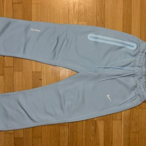 Nike NOCTA ljusblå mjukisbyxor - Helt ny, oanvänd. Snygga ljusblå mjukisbyxor från Nike NOCTA med elastisk midja och dragsko, diskret NOCTA- och Nike-logga samt coola detaljer som dragkedjeficka och tryck på benen. Perfekta för chill eller streetwear. Mjukt material som känns skönt hela dagen. Ingen prislapp eller liknande.