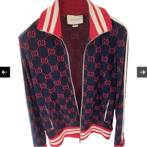 Gucci bomberjacka med GG-mönster - Snygg bomberjacka från Gucci i marinblått med rött GG-mönster över hela jackan. Jackan har vita och röda ränder på krage, ärmslut och nederkant samt dragkedja framtill. Tillverkad i mjuk polyester och har en sportig vibe med klassisk Gucci-stil. Kvitto finns 