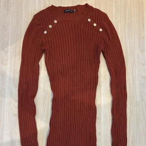 Rostbrun ribbad klänning Brave Soul - Rostbrun ribbstickad klänning från Brave Soul med lång ärm och tight passform. Klänningen har dekorativa silvriga knappar på axlarna och rund halsringning. Perfekt för dig som gillar en enkel men snygg look.