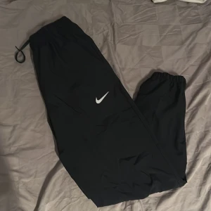 Svarta Nike Dri-FIT träningsbyxor - Svarta träningsbyxor från Nike med Dri-FIT-teknologi. Byxorna har elastisk midja med snörning och en vit Nike-logga på benet. Perfekta för träning eller chill, tillverkade i lätt och syntetiskt material som andas.
