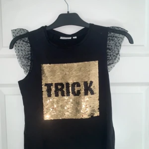 Svart paljettopp KappAhl 146/152 - Cool svart topp från KappAhl med vändbara paljetter framtill som visar texten 'TRICK' eller 'TREAT' i guld och svart. Puffiga ärmlösa axlar i transparent mesh med svarta prickar ger en extra festlig touch. Perfekt för halloween eller när du vill sticka ut.