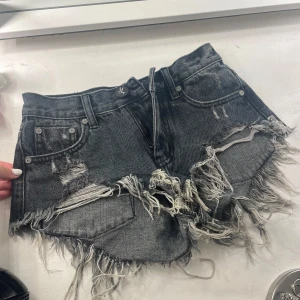 Svarta jeansshorts från One Teaspoon - Riktigt snygga svarta jeansshorts från One Teaspoon med extremt fransig och rå kant, slitna detaljer och låg midja. Klassisk femficksmodell med dragkedja och knapp framtill. Perfekta för en edgy streetstyle-look.