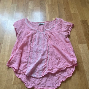 Rosa broderad blus från Svea - Supersöt rosa blus från Svea i luftig bomull med vita broderier framtill och längs halsringningen. Blusen har korta ärmar, rundad nederkant och en avslappnad passform. Perfekt för dig som gillar romantiska detaljer och en chill vibe.