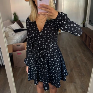 Klänning med blommor  - Svart klänning med vita blommor, v-ringad hals och långa ärmar. Klänningen har knytband i midjan samt dragkedja i ryggen.  Är från Glamourus UK men köpt på Asos. Står ej vad det är för storlek men skulle säga större xs/mindre S