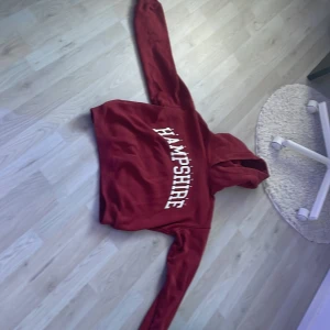 Röd hoodie Hampshire H&M XS - Snygg röd hoodie från H&M i storlek XS med vit text 'HAMPSHIRE' på bröstet. Klassisk modell med huva och långa ärmar, perfekt för chill dagar. Mjuk insida och skön passform. Passar dig som gillar enkel och stilren streetwear.
