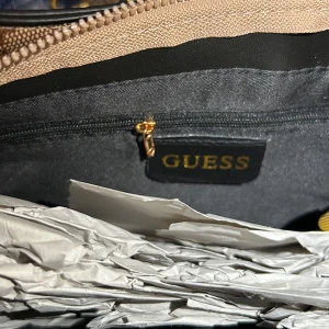 Beige axelväska  - Säljer en snygg beige axelväska från Guess med brunt logomönster över hela väskan. Väskan har guldfärgade detaljer, kedja och G-spänne framtill. Tillverkad i syntetmaterial och har dragkedja upptill. Perfekt för dig som vill ha en trendig och stilren accessoar.
