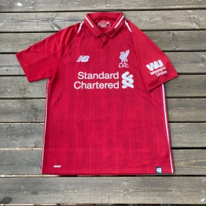 Liverpool FC röd matchtröja New Balance - Liverpool FCs officiella matchtröja från New Balance i rött med vita detaljer. Tröjan har klassisk krage, korta ärmar och klubbmärke samt sponsorloggor på bröst och ärm. Tillverkad i lätt och ventilerande polyester, perfekt för fotboll eller supporterliv.