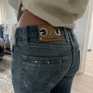 Vintage Dolce & Gabanna lowwaist bootcut jeans - Snygga blå jeans från Dolce & Gabbana med bootcut passform och låg midja. Jeansen har slitna detaljer, nitar på bakfickorna och klassisk D&G-lapp i skinn baktill. Perfekta för dig som gillar en cool och avslappnad stil.