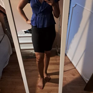 Blå omlottopp med knytband - Snygg blå omlottopp med korta ärmar och v-ringning. Toppen har ett knytband i midjan som ger en fin form och volangkant nertill. Perfekt för dig som gillar en feminin och trendig stil.