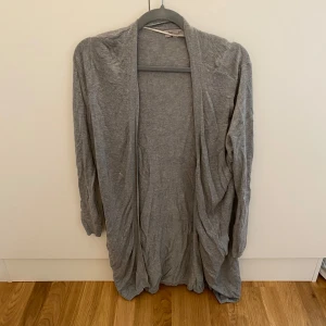 Grå lång kofta  - Lång, öppen kofta. Perfekt att slänga på sig över en t-shirt eller linne. Koftan har långa ärmar och en avslappnad passform som ger en chill vibe.