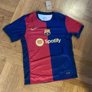 FC Barcelona Nike fotbollströja S - Snygg FC Barcelona matchtröja från Nike i storlek S. Tröjan har klassiska blå och röda fält, klubbmärke på bröstet och guldfärgad Nike-logga. Spotify-logga på framsidan. Tillverkad i lätt och ventilerande material, perfekt för fotbollsplanen.