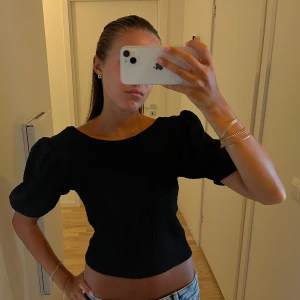 Svart topp med puffärmar - Svart elegant topp med korta puffärmar och rundad halsringning. Modellen är croppad och har en smockad, stretchig struktur som sitter snyggt på kroppen. Toppen har en djupare urigning i ryggen.