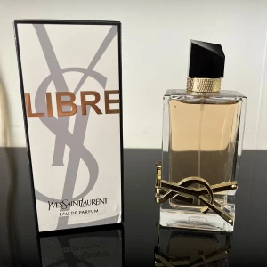 Yves Saint Laurent Libre EdP 90ml - YSL Libre Eau de Parfum i en lyxig, rektangulär glasflaska med guldiga detaljer och svart asymmetrisk kork. Parfymen har en ljus bärnstensfärgad vätska och kommer i en elegant vit och svart kartong med stora bokstäver och YSL-logga.