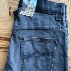 Grå Nudie Jeans Thin Finn  - Säljer ett par gråa Nudie Jeans Thin Finn med skinny fit och låg midja. Klassisk femficksmodell med svarta detaljer och broderad bakficka. Perfekta för dig som gillar smal passform och stilren look.
