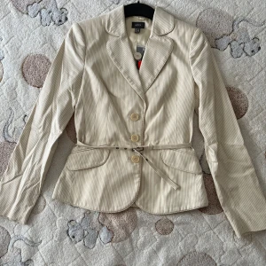 Beige randig kavaj från Mexx - Snygg beige kavaj från Mexx med tunna ränder, klassisk krage och tre knappar framtill. Jackan har ett smalt bälte i midjan och två fickor med lock. Perfekt för en stilren look och passar till många outfits.