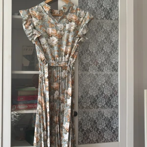 Lång blommig klänning med volangärm - Superfin långklänning med ljusblå botten och stora blommor i vitt, beige och orange. Klänningen har volangärmar, v-ringning med knapp och plisserad kjol. Perfekt för sommarens alla tillfällen när du vill känna dig extra söt och bekväm.