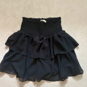 Svart volangkjol från Chelsea XS - Supersöt svart kortkjol från Chelsea med bred smock i midjan och dubbla volanger. Kjolen är luftig och har en flowy känsla, perfekt för att skapa en cool och avslappnad look. Passar dig som gillar feminina detaljer och vill ha något som sticker ut.