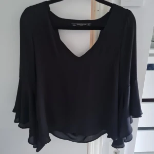 Svart blus med volangärm från Zara - Supersnygg svart blus från Zara Trafaluc med v-ringning och öppen rygg. Blusen har långa, vida volangärmar och är gjord i ett lätt, flowy material som ger en riktigt cool siluett. Perfekt för dig som gillar statement-ärmar och stilren look.