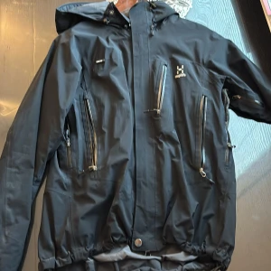 Svart vindjacka från Haglöfs med huva - Snygg svart vindjacka från Haglöfs med flera dragkedjeförsedda fickor och justerbar huva. Jackan har en sportig look och är perfekt för utomhusaktiviteter. Materialet är slitstarkt och vattenavvisande, med detaljer som dragsko i midjan och logga på bröstet.