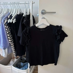 Svart topp med volangärmar från Zara - Svart topp från Zara med korta ärmar och snygga volangdetaljer på axlarna. Enkel men ändå trendig, perfekt att matcha med jeans eller kjol. Mjuk bomullskänsla och rund halsringning.