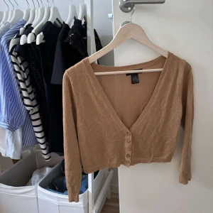 Beige glittrig croppad kofta med knappar - Snygg beige glittrig croppad kofta från ellos med v-ringning och långa ärmar. Koftan har en glittrig finish och stängs med fem knappar framtill. Perfekt att styla med höga jeans eller kjol för en trendig look. Passar till festliga tillfällen.