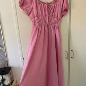 Rosa långklänning med puffärm - Supersöt rosa långklänning med smockad midja, puffiga korta ärmar och knytning vid halsringningen. Klänningen har en luftig och flowy passform, perfekt för varma dagar. Materialet känns lätt och bekvämt mot huden. Jag står inte för retur och för postens slarv.