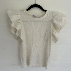 Vit ribbad topp med volangärm Zara - Superfin vit ribbad topp från Zara med breda volangärmar som ger en extra cool touch. Toppen är i mjuk bomull och har en rund halsringning. Perfekt för dig som vill ha något stilrent men ändå unikt i garderoben.