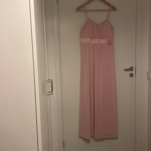 Ljusrosa långklänning med blomdetalj - Romantisk ljusrosa långklänning med smala justerbara axelband och en söt blomdetalj i midjan. Silikonkat runt om för att kunna använda klänningen utan axelband. Klänningen har ett mjukt och lätt, fallande tyg och markerad midja med band. Perfekt för dig som vill ha en feminin och elegant look. Ganska stor i storleken och vid bysten. Använd en gång.