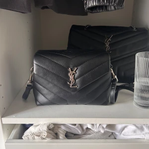 Svart quiltad axelväska från Saint Laurent - Säljer en svart quiltad axelväska från Saint Laurent med ikoniskt YSL-emblem i silver framtill. Väskan är tillverkad i skinn och har en stilren, klassisk design med justerbar axelrem. Perfekt för dig som vill ha en lyxig och tidlös accessoar.