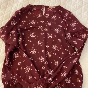 Vinröd blus med blommor i storlek 158 - Vinröd blus med blommor. Blusen har volanger i mitten och nedre delen av ärmarna. Pris går att diskutera