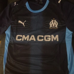 Marseille borta tröja 2025-26 - Snygg bOlympique de Marseille borta fotbollströja i mörkblått med ljusblå detaljer på sidorna och kragen. Tryckt klubbmärke och sponsorlogga på bröstet. Tillverkad i lätt och ventilerande funktionsmaterial, perfekt för träning eller match. Knappt använts och rätt så ny