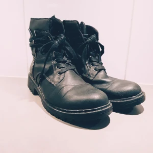 Svarta kängor med snörning strl 37 - Tuffa svarta kängor i skinn med grov sula och snörning framtill. Snygga detaljer med synliga sömmar och metallnitar på sidan. Perfekta för dig som gillar en edgy stil och vill ha skor som funkar hela hösten och vintern.  Ena hällan vid skosnörena har gått sönder (se bild) men inget som stör funktionen & en liten reva fram men inga hål