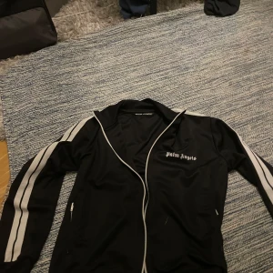 Palm Angels svart vindjacka med ränder - Svart vindjacka från Palm Angels med vita ränder längs ärmarna och dragkedja framtill. Jackan har klassisk sportig look, ståkrage och logga på bröstet. Tillverkad i Italien och gjord i polyester. Perfekt för dig som gillar streetwear vibes.